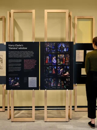 A woman standing reading the Harry Clarke’s 'Geneva' window exhibition panel. The panels feature comprehensive text about the subject and a large digital reproduction of the glass window.