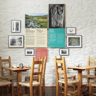 A small exhibit within the café featuring introductory panels about the village and the museum accompanied by framed photographs on a white brick wall surrounded by wooden tables and chairs.