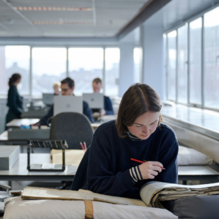 A woman studies an open census book. She holds a red pencil as she examines the pages. In the background, several other people are working at desks with laptops.