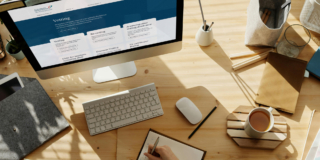 A desk workspace with a monitor showing the Teaching Council website and a person writing in a notebook.