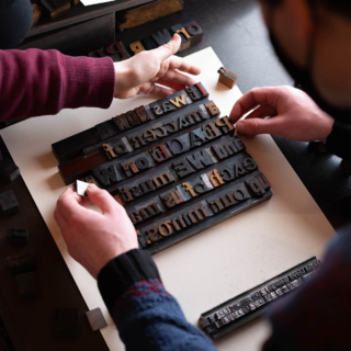 Two individuals carefully arranging wood type blocks on a flat surface, to form a quote. Various other blocks and materials are scattered around.