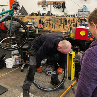 A scene inside a bike repair shop. A man is working on a bicycle wheel, which he is holding in his hands. He is wearing orange gloves and is bent over, focusing on the wheel.