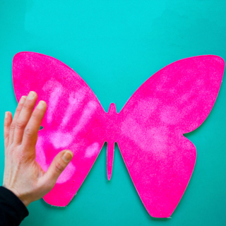 A fluorescent pink cut out of a butterfly with handprints on each wing. The butterfly is fixed to a teal wall. There is a persons hand in the foreground.