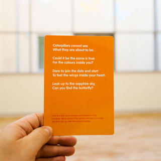 Closeup of an orange card in someones hand. The card says Catterpillars cannot see What they are about to be. Could it be the same is true For the colours inside you? Dare to join the dots and start To feel the wings inside your heart. Look up to the sapphire sky, Can you find the butterfly? Find the dots and numbers somewhere in the building. Move around the space until you can make the dots join up with the picture nearby.