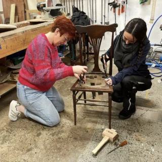 Two woman within a workshop either side of an old chair. They are working together using various tools and are in the process of refurbishing the chair.