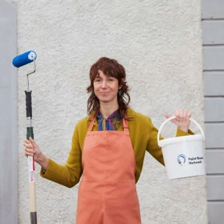 A woman standing in front of a textured wall, holding a paint roller with a blue roller cover in one hand and a white paint bucket in the other. She is wearing an orange apron over a greenish-yellow sweater and has a friendly expression with a slight smile. The paint bucket has a logo on it that reads 
