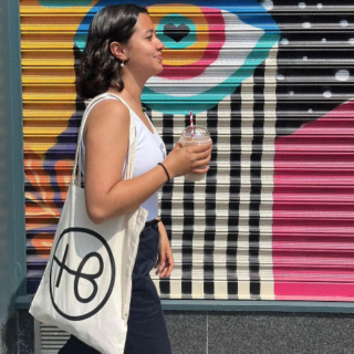 A woman wearing a Hopeless Botanics tote bag on her shoulder whilst walking down a street with colourful street art on a shopfront.