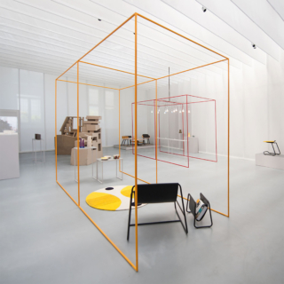 A modern, minimalist exhibition space with a large orange cube structure in the center. Inside the cube are various design objects including furniture, lighting, and books. Red lines outline additional display areas within the space.