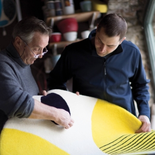 Two men from Ceadogan Rugs examine a rug with bold abstract patterns in yellow, black, and white. One wears a gray sweater with glasses, the other a navy jacket. They are in a workshop with shelves holding various materials.
