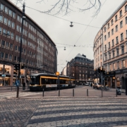 A tram in the centre of Helsinki, Uusimaa, Finland. The road is cobbled and there are large curved brown bricked buildings behind the tram.