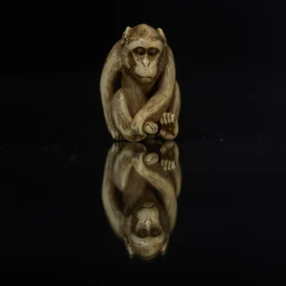 Carved ivory Netsuke in the shape of a monkey, with detailed features, sitting in a reflective pose on a dark surface, creating a mirrored reflection.