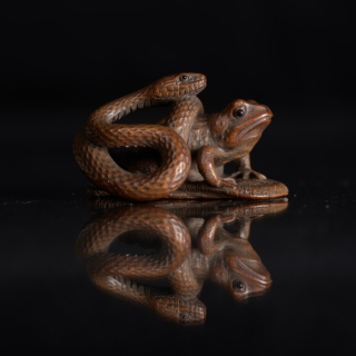Wooden Netsuke carving depicting a snake coiled around a frog, with intricate texture details, set against a reflective dark surface.