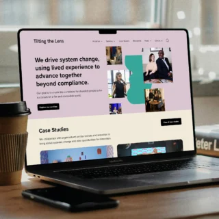 A laptop on a desk featuring the homepage of Tilting the Lens website with a mission statement that reads: 'We drive system change, using lived experience to advance together beyond compliance.' The site features a navigation menu with sections including About Us, Services, Case Studies, Resources, News, and Contact Us. Below the mission statement is a case studies section.