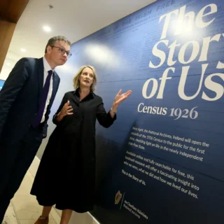 Two people stand outside the National Archives of Ireland building beside large window graphics promoting ‘The Story of Us – Census 1926’ exhibition