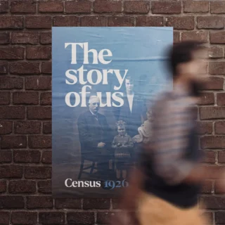 Census 1926 street poster reading 'The Story of Us', featuring a black and white family portrait from the 1920s, wheat-pasted on a brick wall with a pedestrian passing in the foreground.