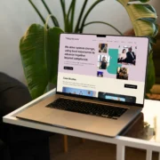 A laptop on a glass coffee table featuring the homepage of Tilting the Lens website with a mission statement that reads: 'We drive system change, using lived experience to advance together beyond compliance.' The site features a navigation menu with sections including About Us, Services, Case Studies, Resources, News, and Contact Us. Below the mission statement is a case studies section.